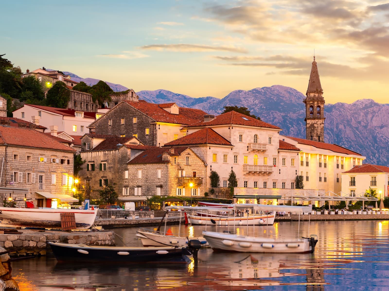 Abendstimmung in Perast an der Bucht von Kotor: historische Steinhäuser mit roten Dächern, Kirchturm und Boote am Ufer vor Bergen im Sonnenuntergang