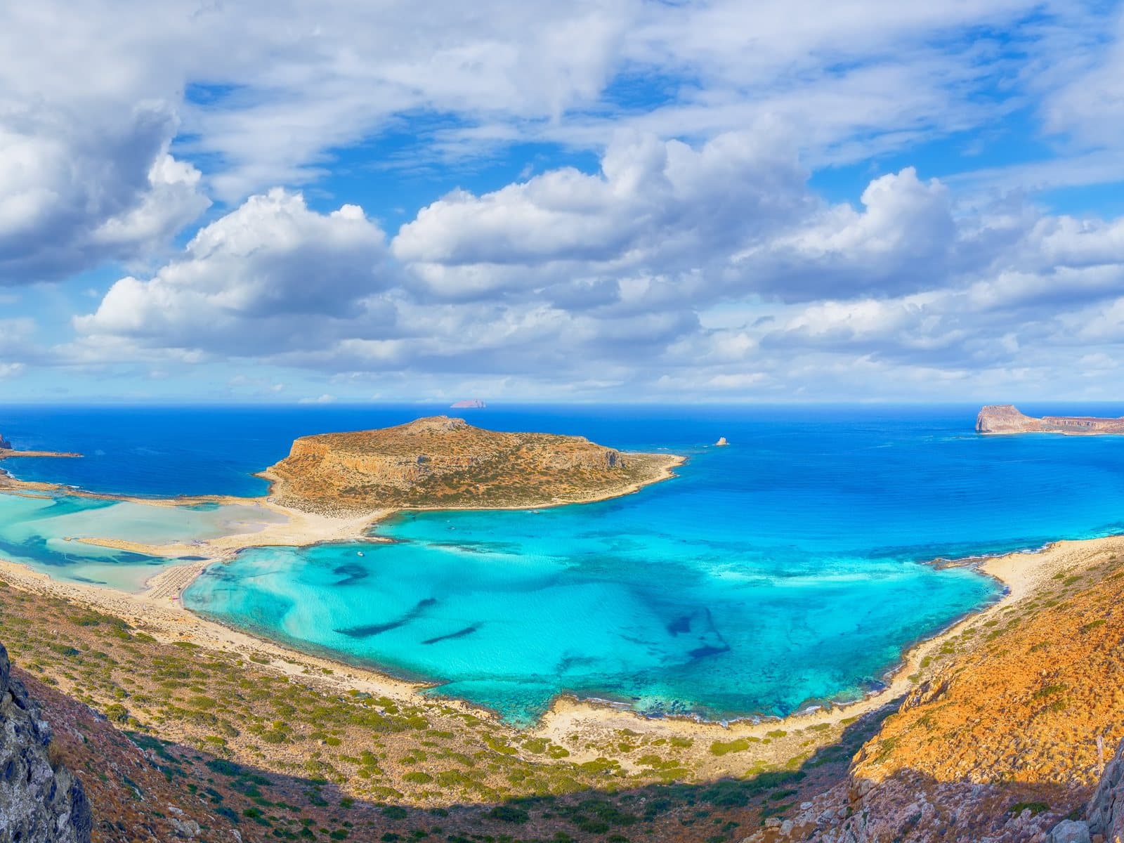Lagune von Balos auf Kreta: türkisfarbenes, flaches Wasser und helle Sandbänke zwischen felsigen Hügeln, dahinter das tiefblaue Meer und die Insel Gramvousa unter weitem Wolkenhimmel