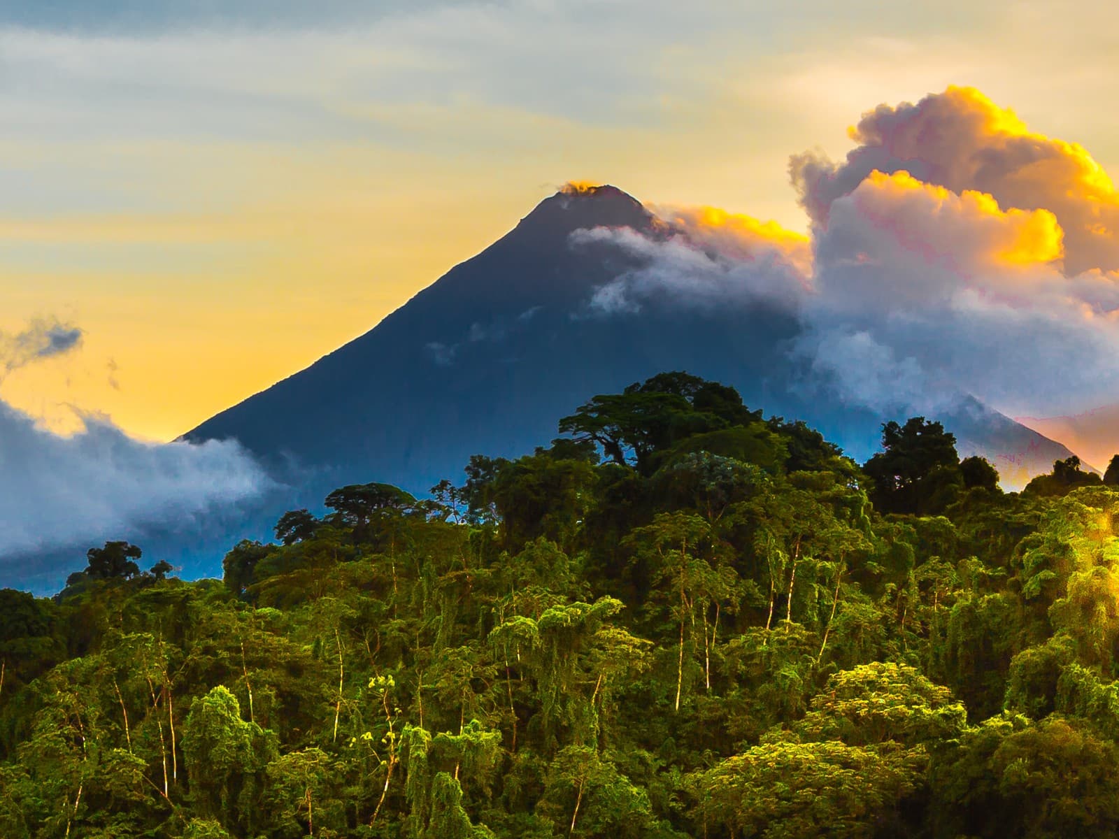 Kegelförmiger Vulkan über dichtem tropischem Regenwald in Costa Rica bei Sonnenuntergang, Wolkenbänder ziehen um den Gipfel, goldener Himmel