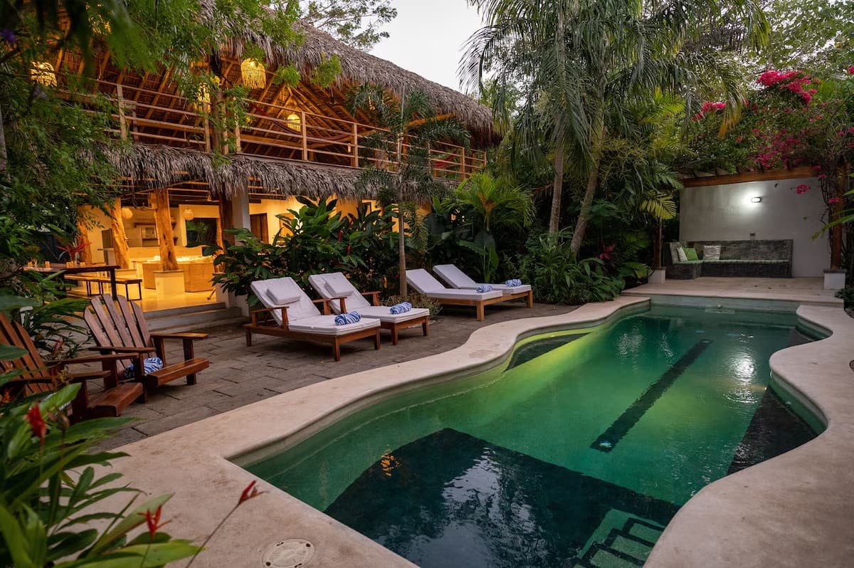 Ferienhaus in Nosara in der Abenddämmerung: beleuchtete Terrasse unter Palmstrohdach, Sonnenliegen und Holzstühle am geschwungenen, grün schimmernden Pool, umgeben von tropischen Pflanzen und pinker Bougainvillea