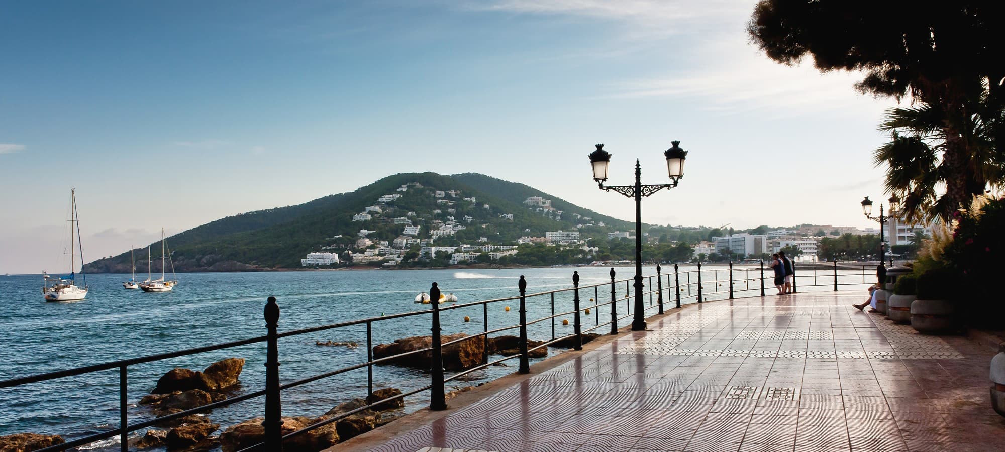 Uferpromenade von Santa Eulalia mit schmiedeeisernem Geländer und Laternen am Meer, im Hintergrund ein grüner Hügel mit weißen Häusern und ankernden Segelbooten
