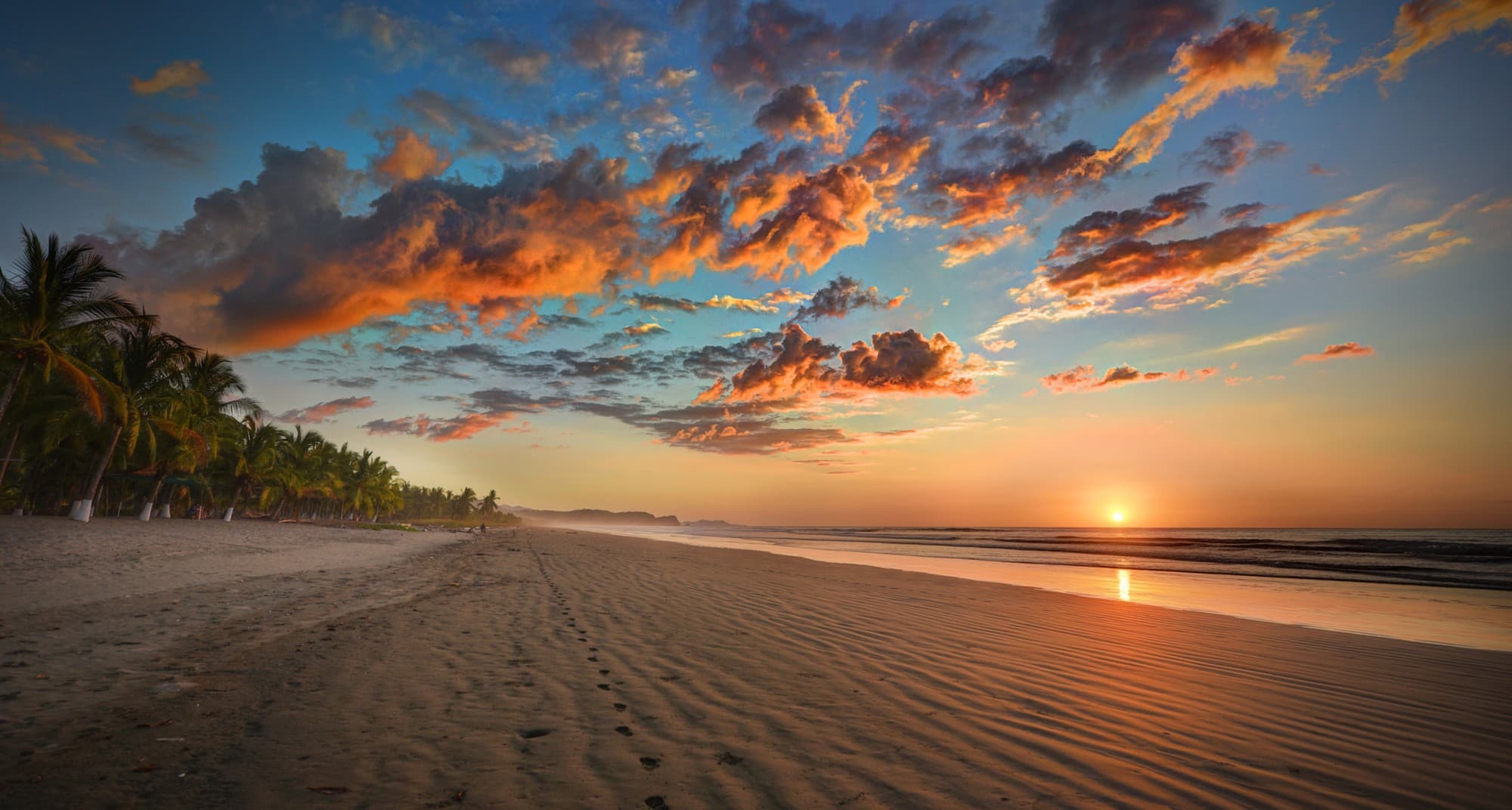 Sonnenuntergang am weiten Sandstrand bei Nosara: orange leuchtende Wolken über dem Pazifik, Palmen am linken Bildrand und Rippelmuster mit Fußspuren im Sand