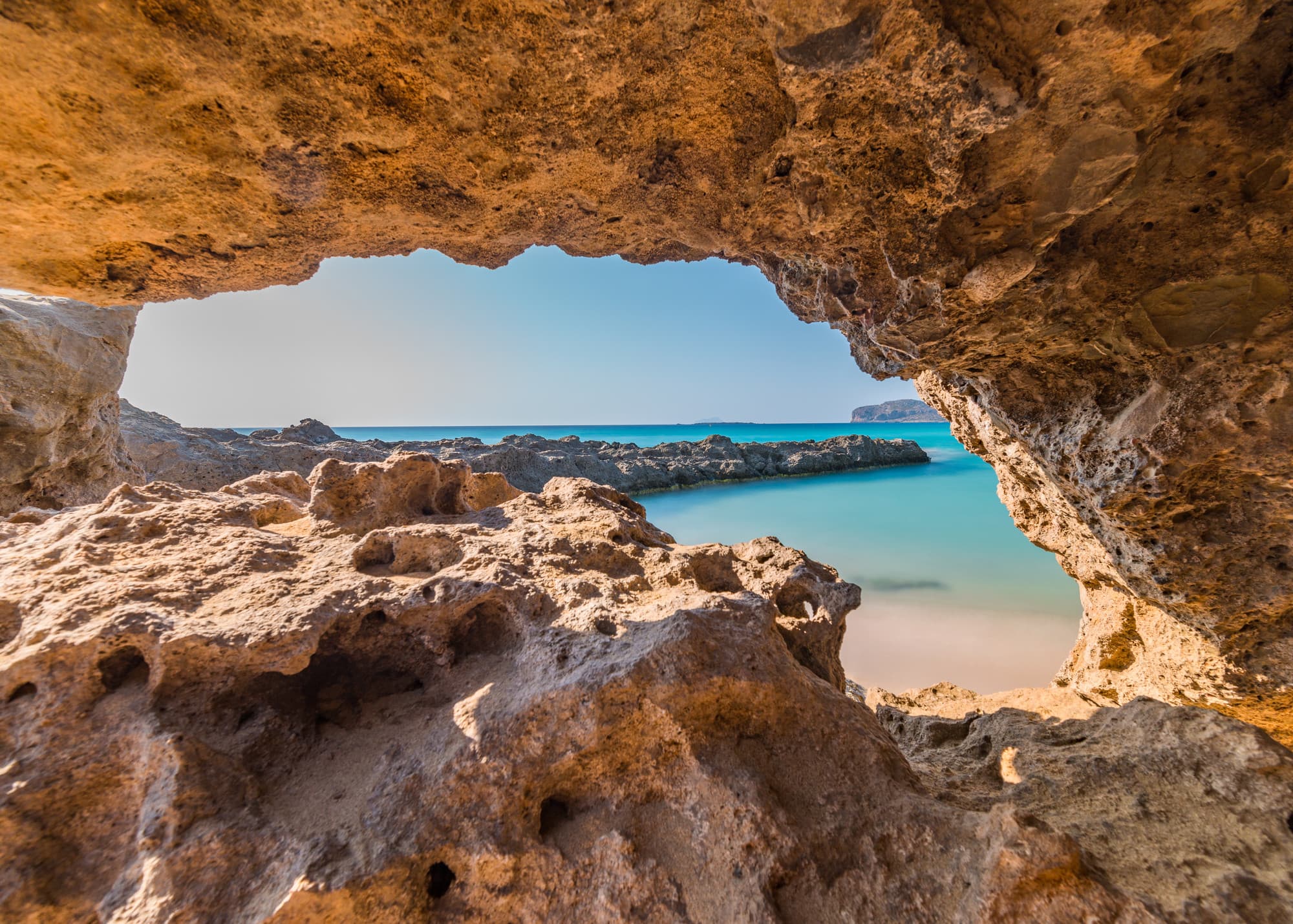 Blick aus einer rötlichen Felsenhöhle auf eine türkisfarbene Bucht mit Sandstrand und Mole