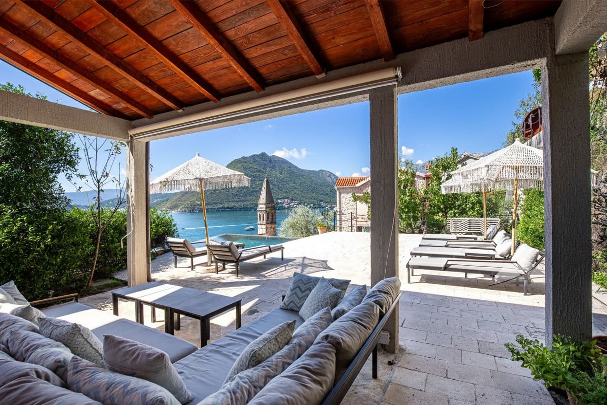 Überdachte Terrasse der Monte Bay Retreat Villa mit Loungesofas, Sonnenschirmen und Liegen am Pool, Blick auf die Bucht von Kotor und den Kirchturm von Perast