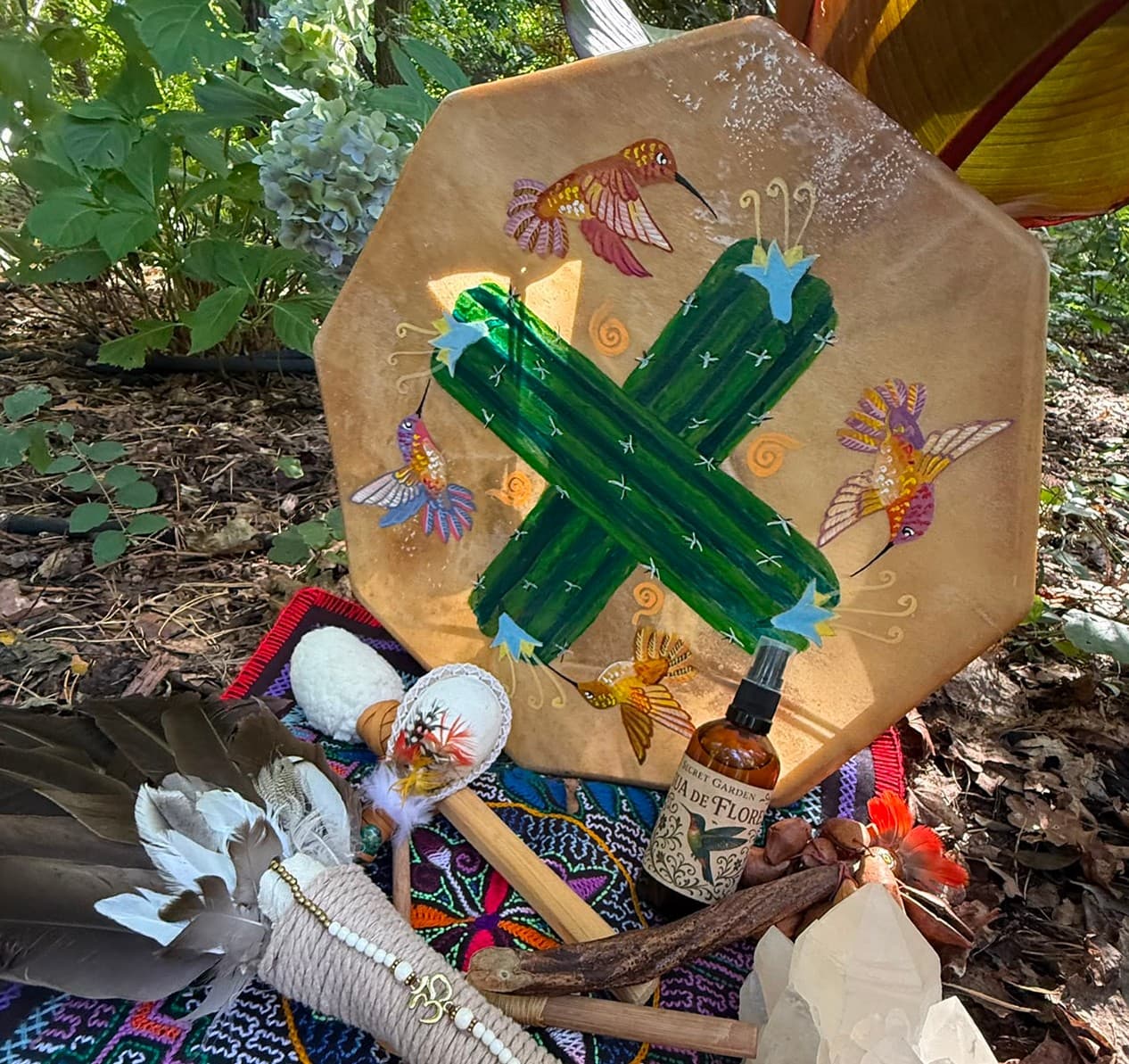 Schamanischer Altar im Dschungel: bemalte achteckige Rahmentrommel mit grünem Kreuz und Kolibris, davor Federfächer, Rasseln, ein kleines Fläschchen und ein Bergkristall auf einem bunten Webtuch