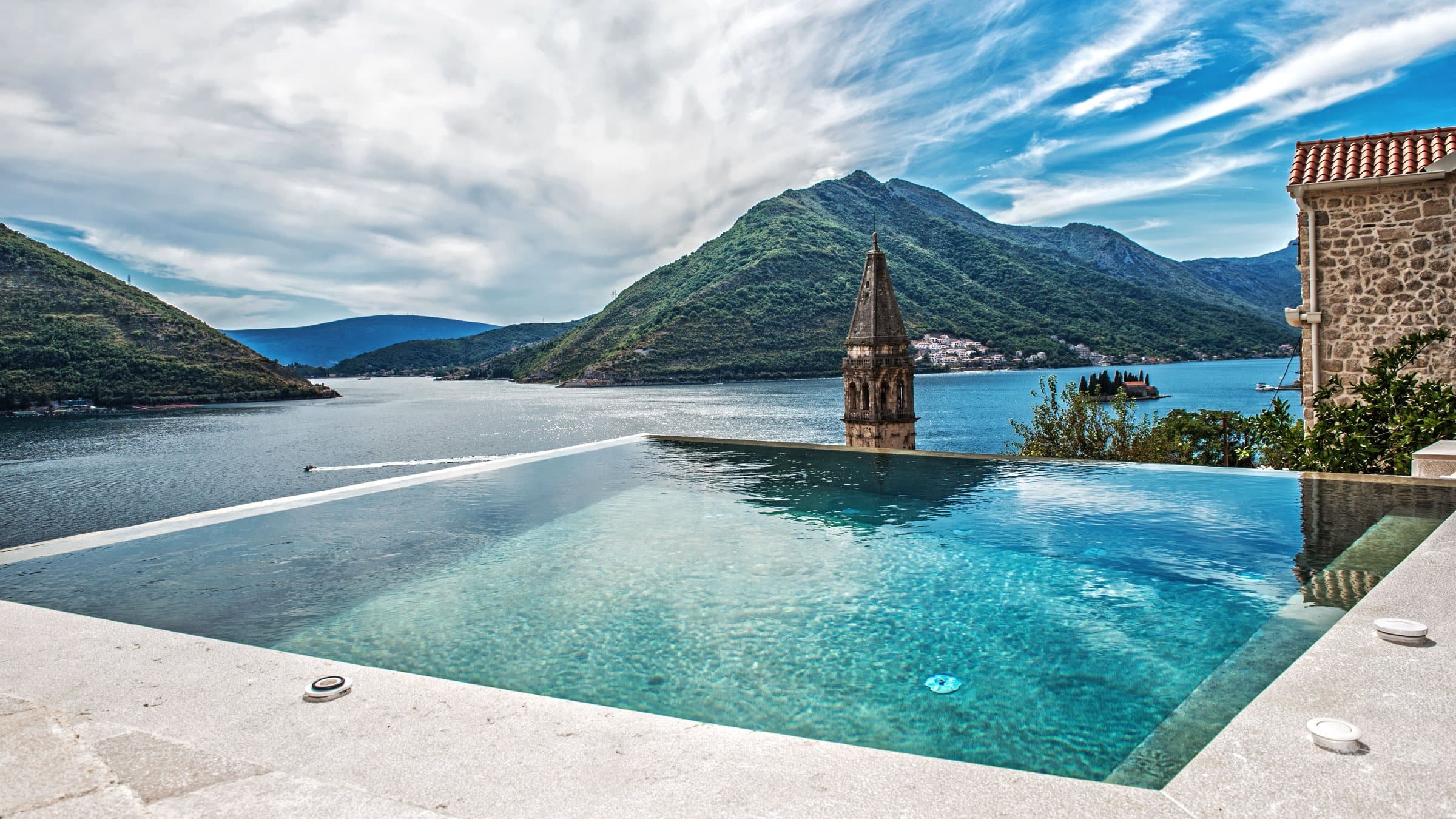 Infinity-Pool auf der Terrasse der Monte Bay Retreat Villa: türkisfarbenes Wasser, dahinter der Kirchturm von Perast, die Bucht von Kotor mit ihren kleinen Inseln und grüne Berge unter weitem Himmel