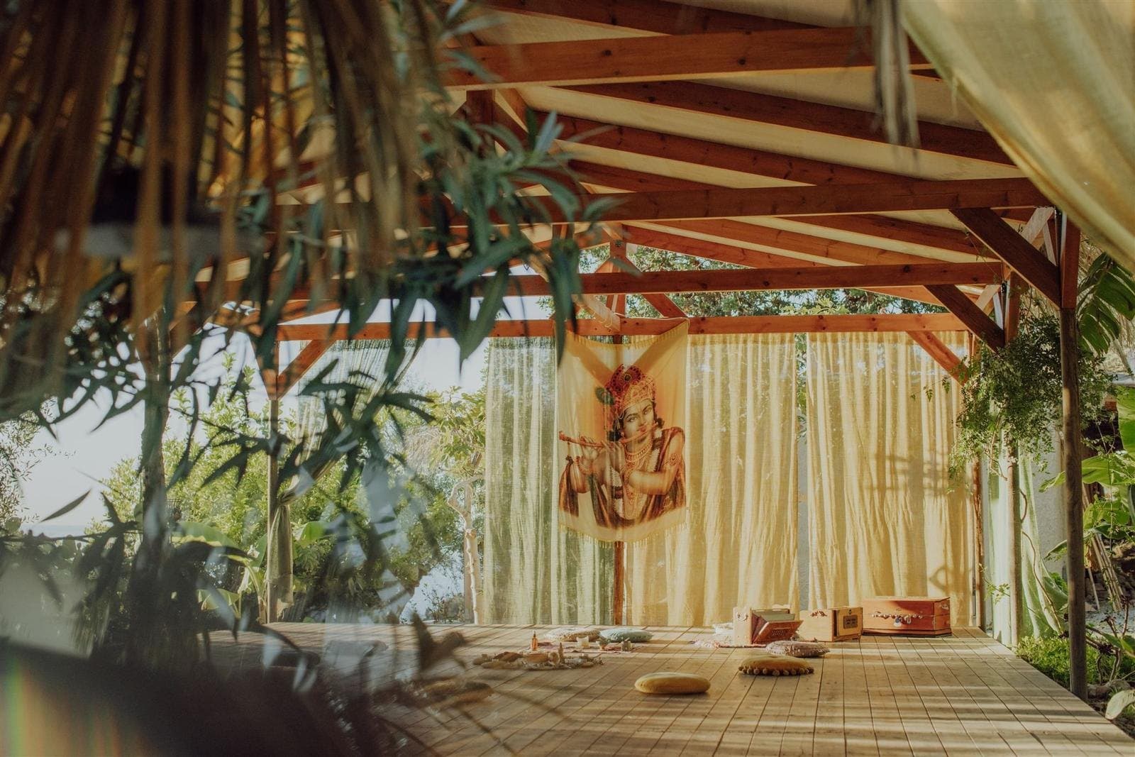 Offener Yoga-Pavillon im Rinido Eco Village mit Holzbalkendach, hellen Vorhängen und einem Wandbehang mit flötespielendem Krishna, Meditationskissen auf dem Holzboden und üppigem Grün ringsum