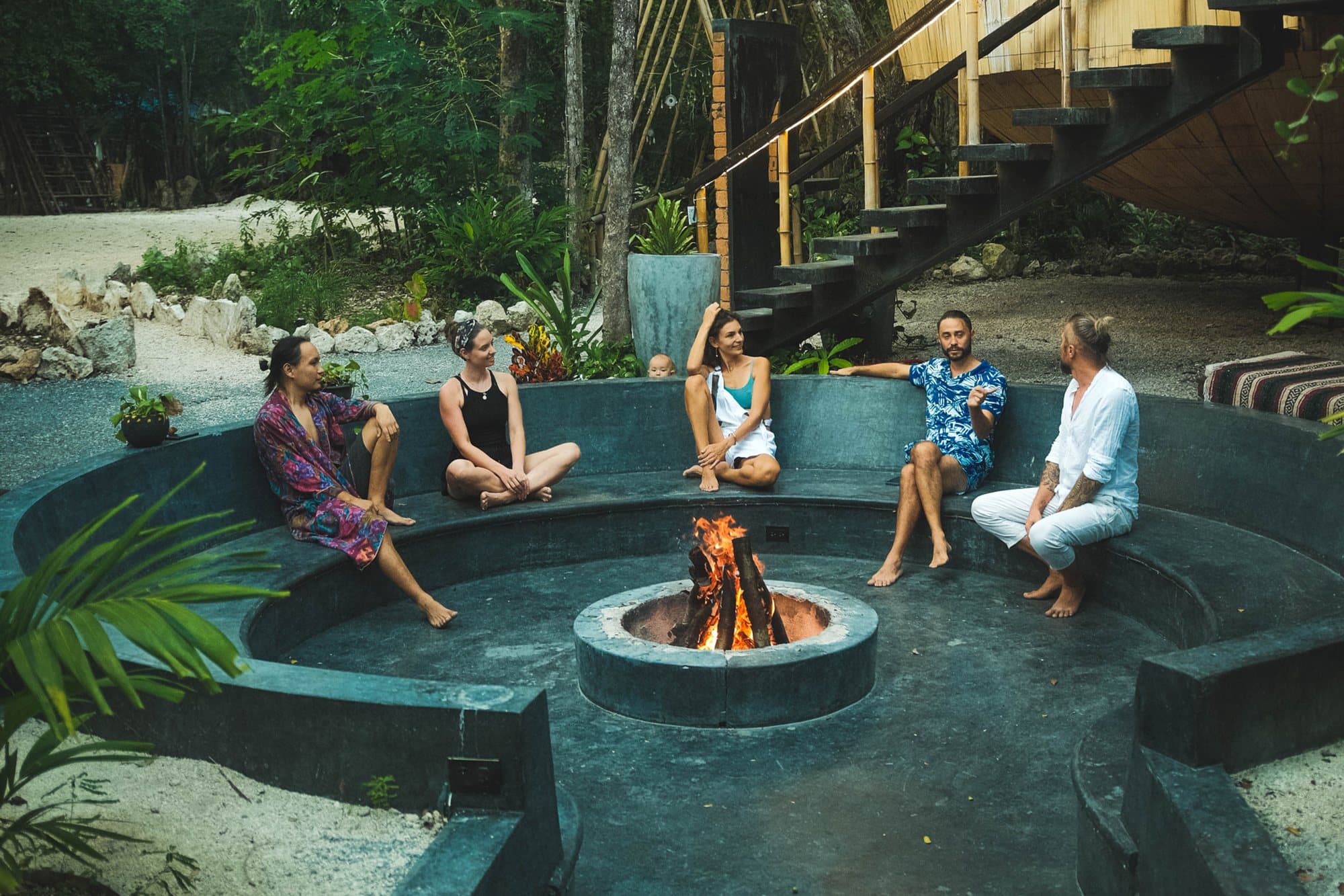 Eine kleine Gruppe sitzt entspannt auf der runden Steinbank einer Feuerstelle im Ekumal Sanctuary Eco Retreat Center, in der Mitte brennt ein Feuer, ringsum Dschungelgrün und eine Holztreppe