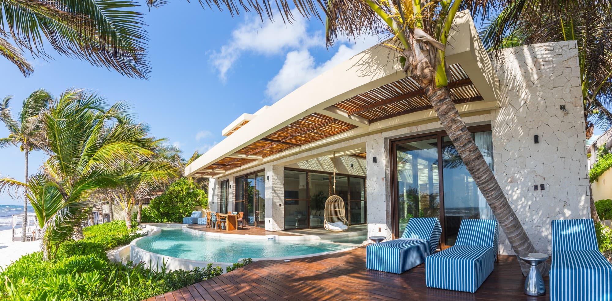 Moderne weiße Villa des Ana y José Hotels in Tulum mit Holzterrasse, Pool, blau-weiß gestreiften Liegen und Palmen unter blauem Himmel
