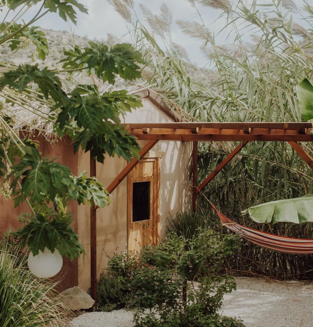 Kleines Tiny House mit Holztür und Pergola, dazu Hängematte, Bananen- und Papayapflanzen, Pampasgras und eine runde Gartenleuchte
