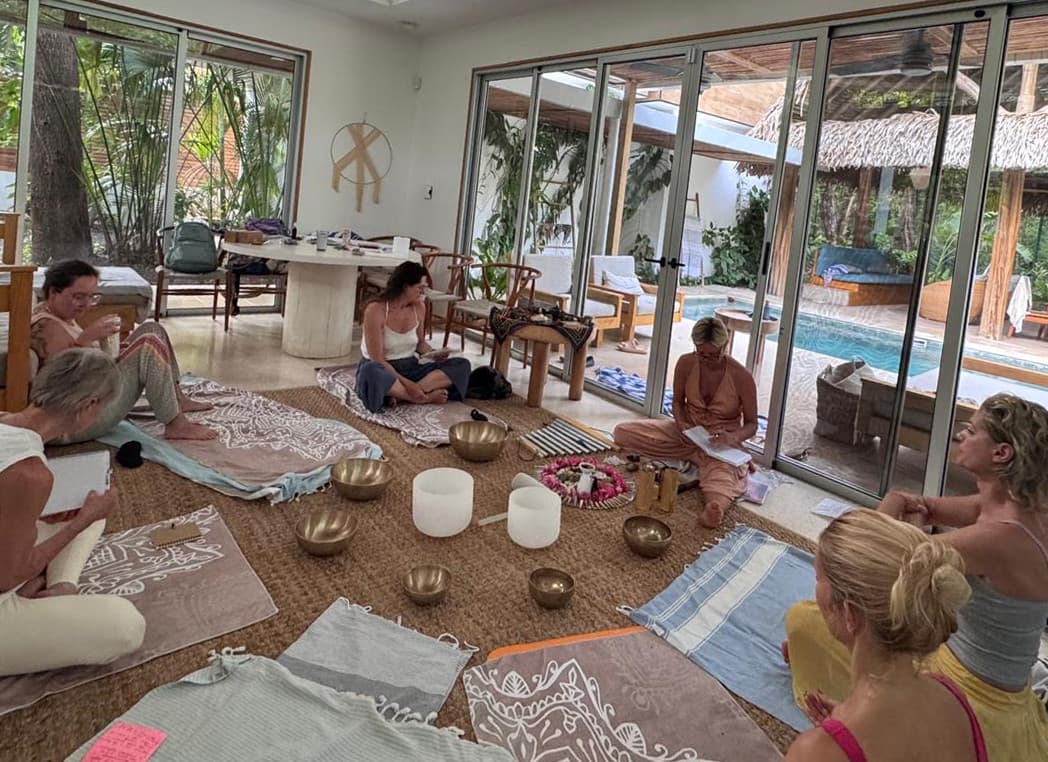 Gruppe sitzt bei einer Klangzeremonie mit Klangschalen im hellen Yogaraum mit Blick auf den Pool